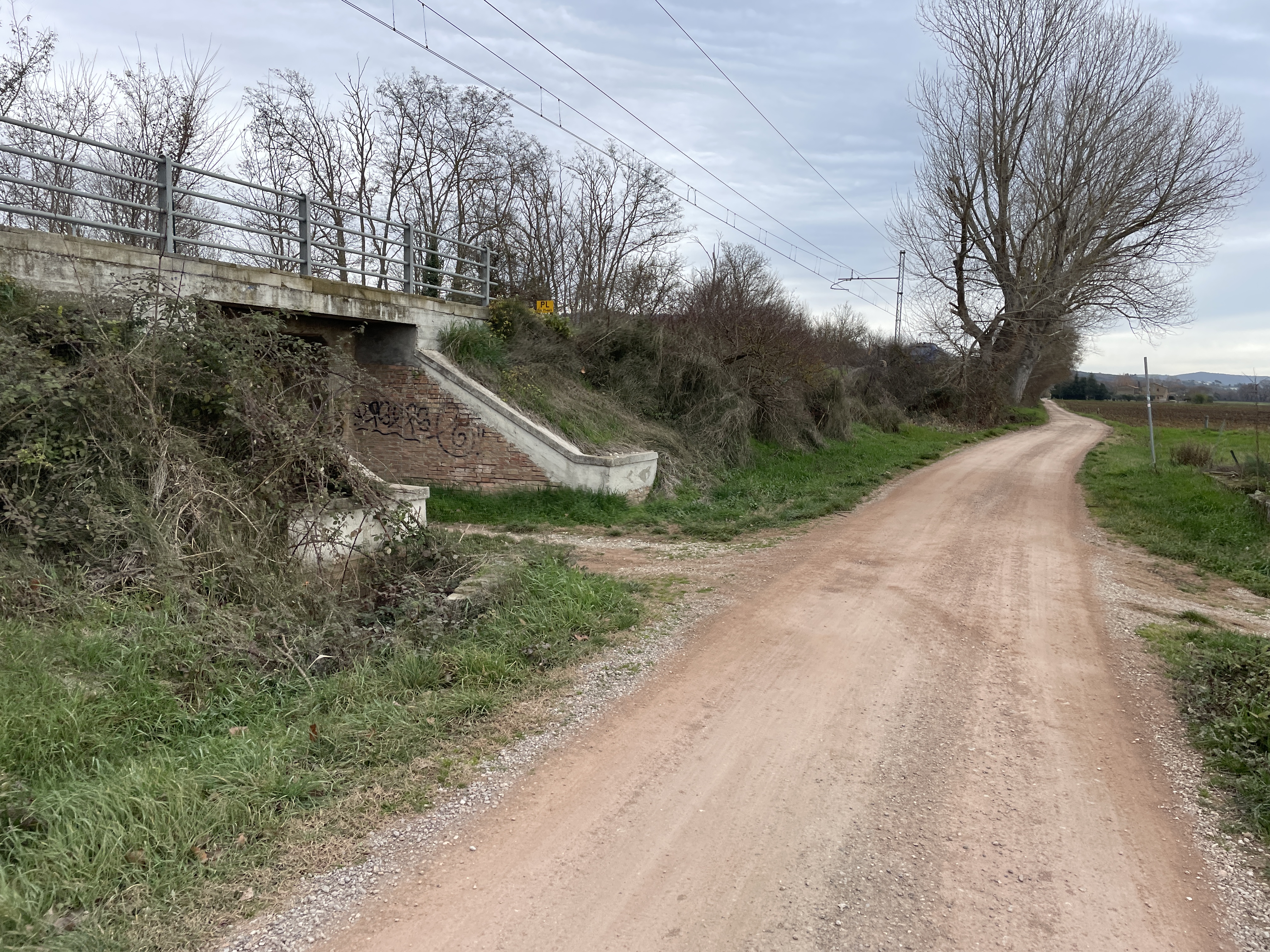 Dirt bikeway next to concrete and brick railroad underpass. Fields in background and elevated railway on right.